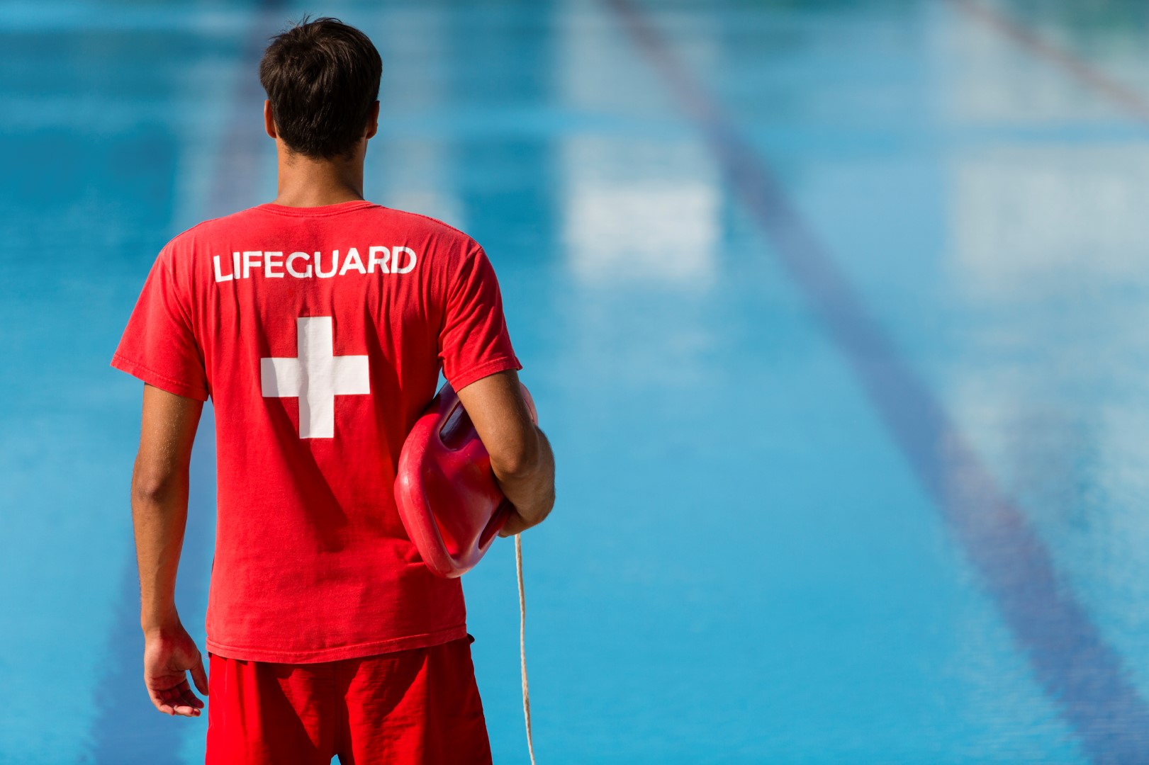 Rear view of male lifeguard with emergency equipment in red uniform watching swimming pool.